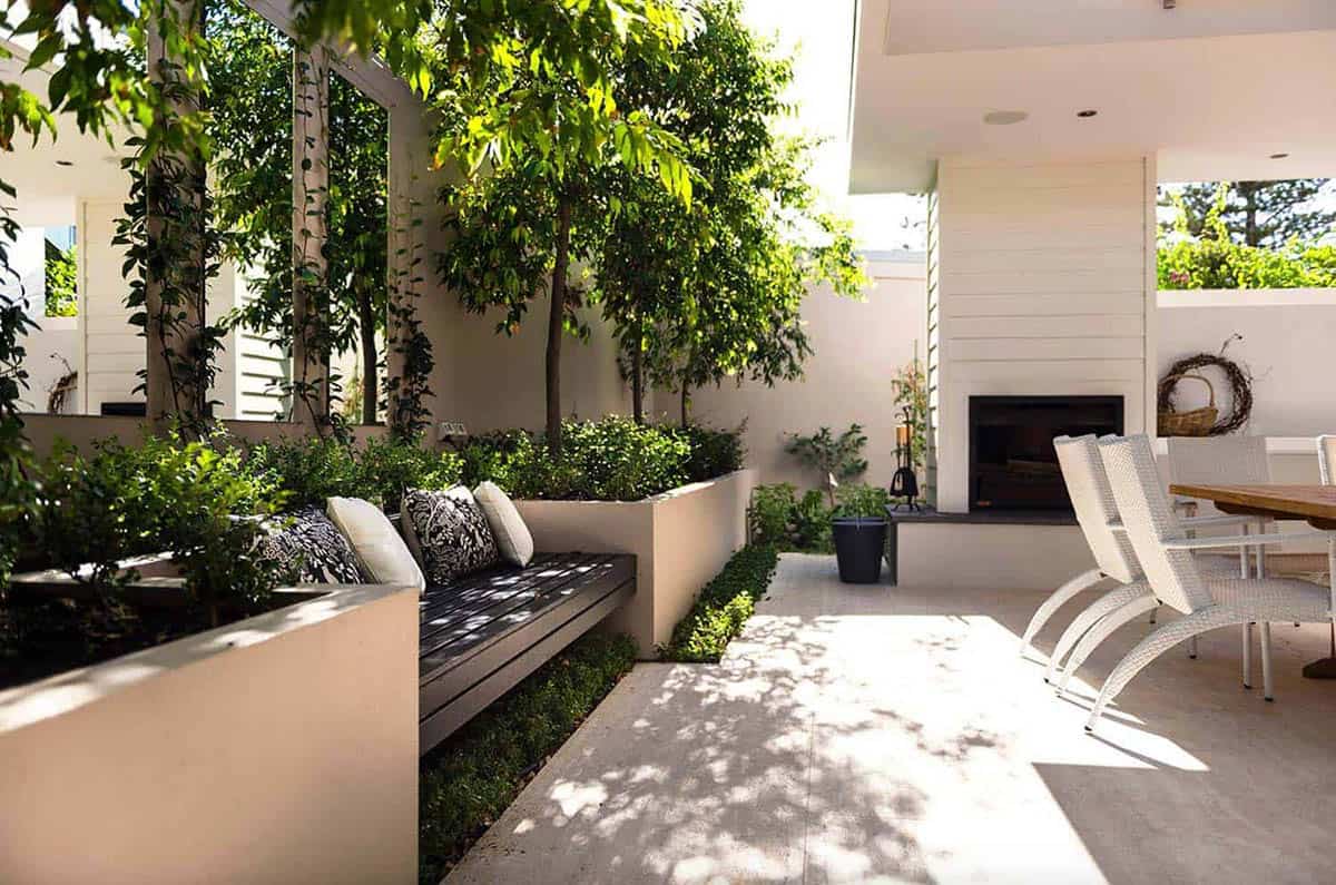 Modern outdoor patio with built-in timber bench, decorative pillows, lush garden trees, white wicker dining chairs, and a wall-mounted fireplace