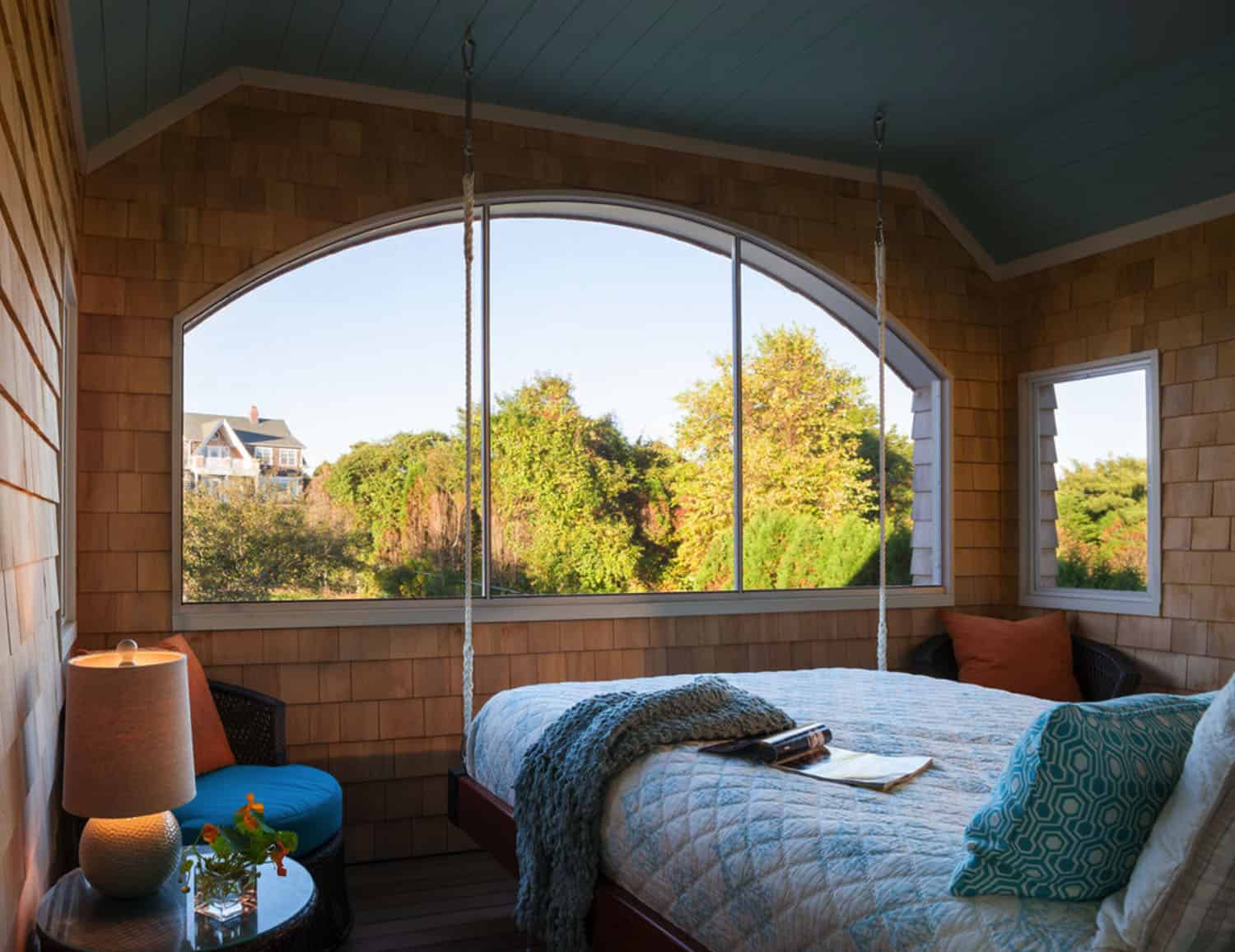bed hanging from ceiling on sleeping porch with blue blanket and large windows with a view of trees