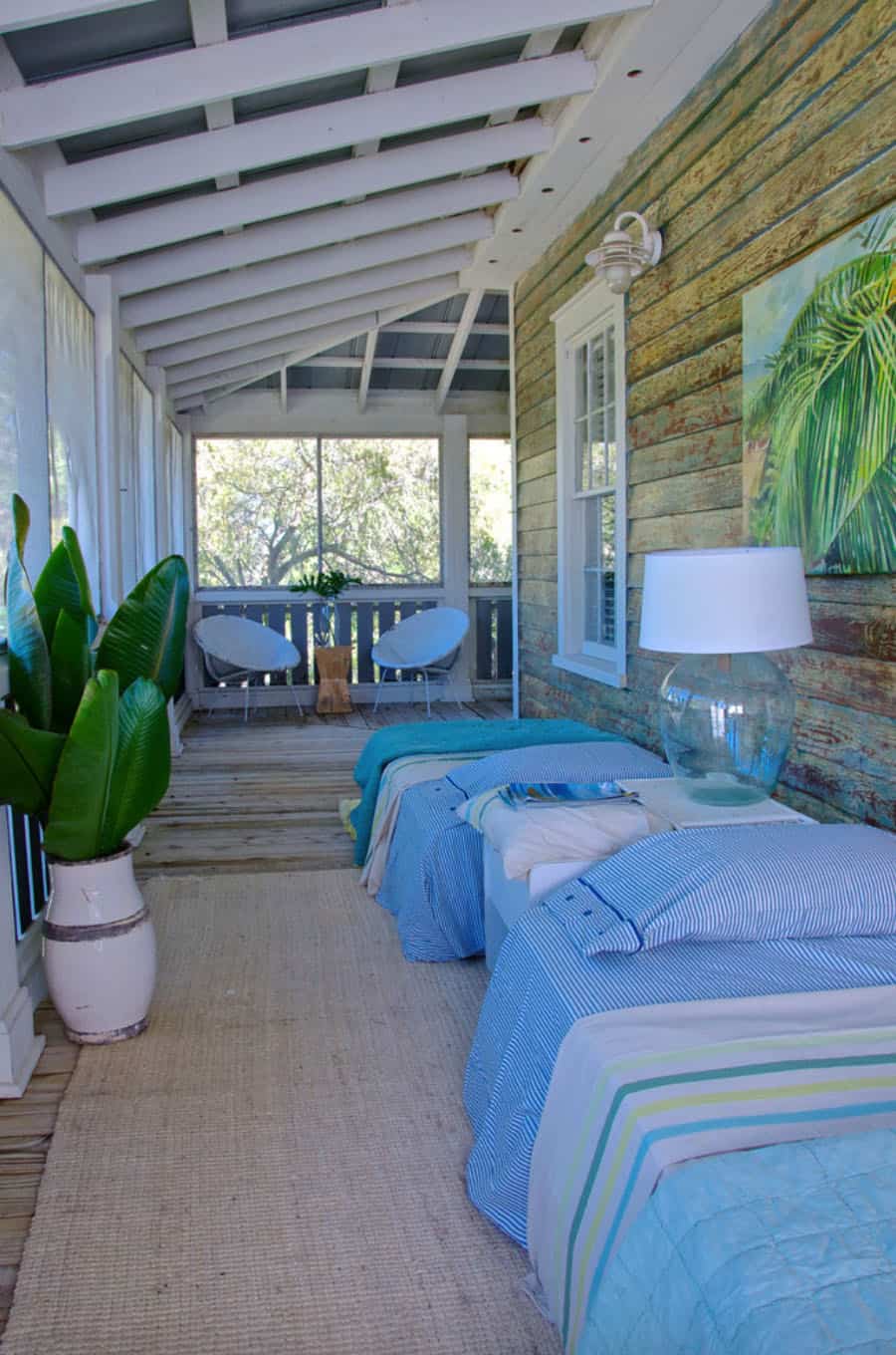 long sleeping porch with two bed with blue striped blanket, large plant and lamp