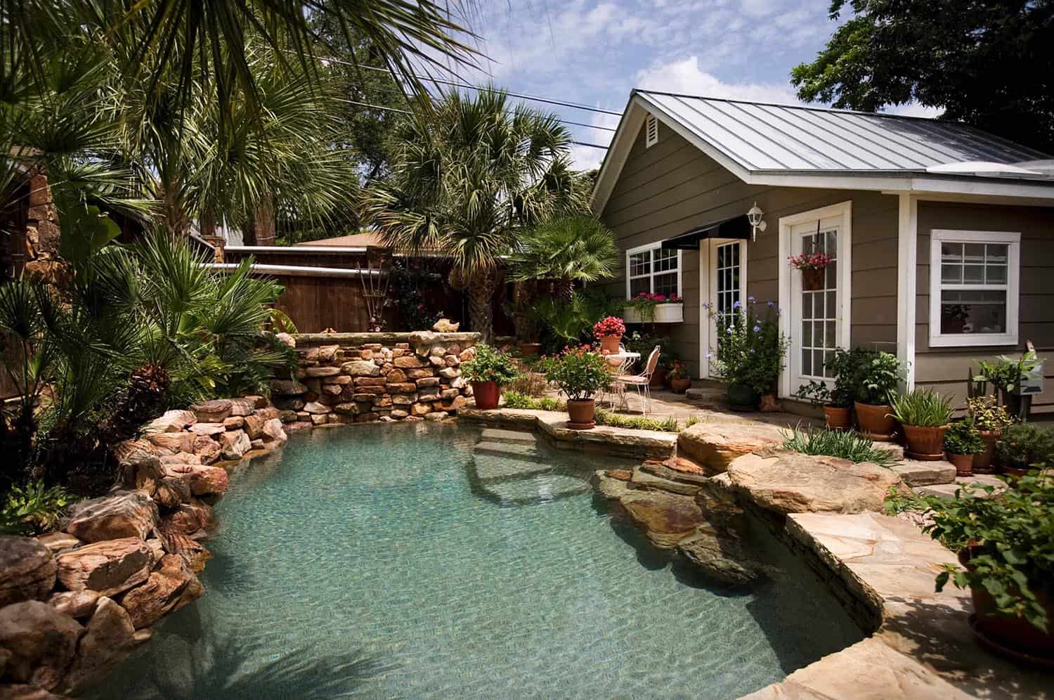 pool with rock formations on the side with trees, a house and plants in the background 