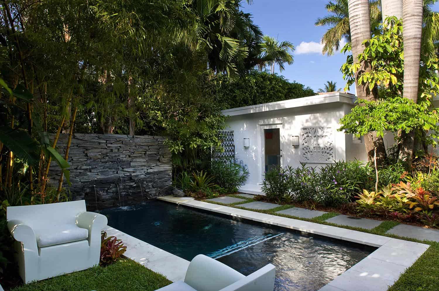 small rectangle pool with rock formation waterfall and sitting area with house in background 