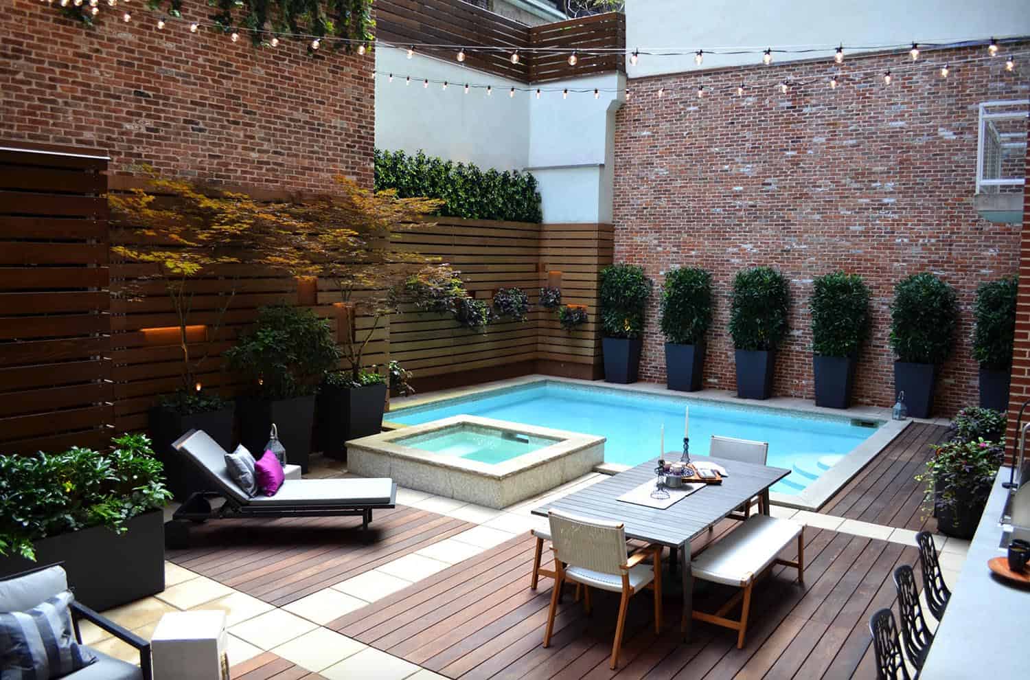 small pool with neutral colored sitting area and plants, bushes and a brick house in the background 