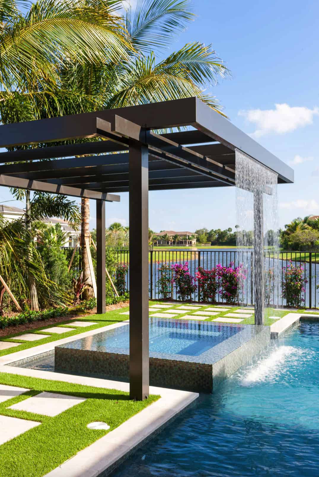 outdoor hot tub with waterfall coming out of pergula and connected to a pool. palm trees and lake are in the background. 