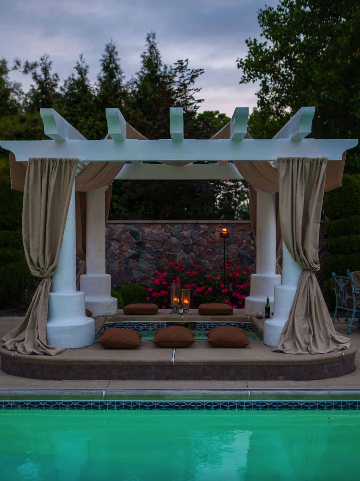 hot tub under a pergola with curtains bunched on the side, rock and flowers in the background 