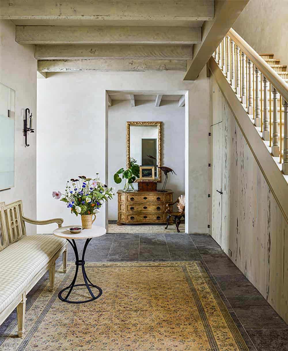 A hallway with a wooden bench, round table with a flower vase, patterned rug, and a staircase; a chest of drawers and mirror are visible in the background