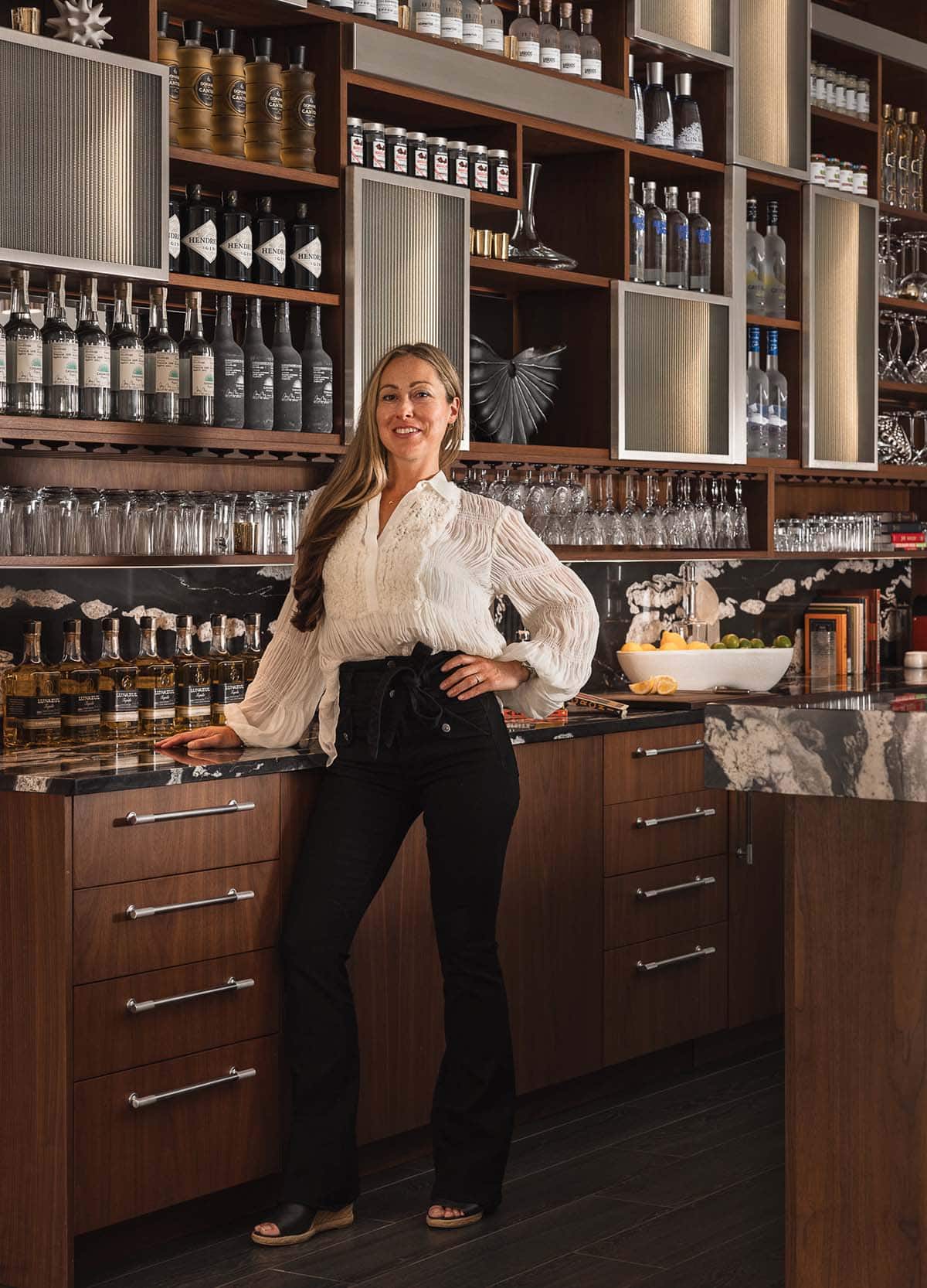 Stylish woman in modern home bar with wood and stone cabinetry, bottles, and glassware
