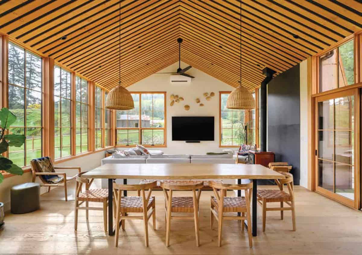 Modern farmhouse interior with vaulted wooden ceiling and large windows overlooking nature.