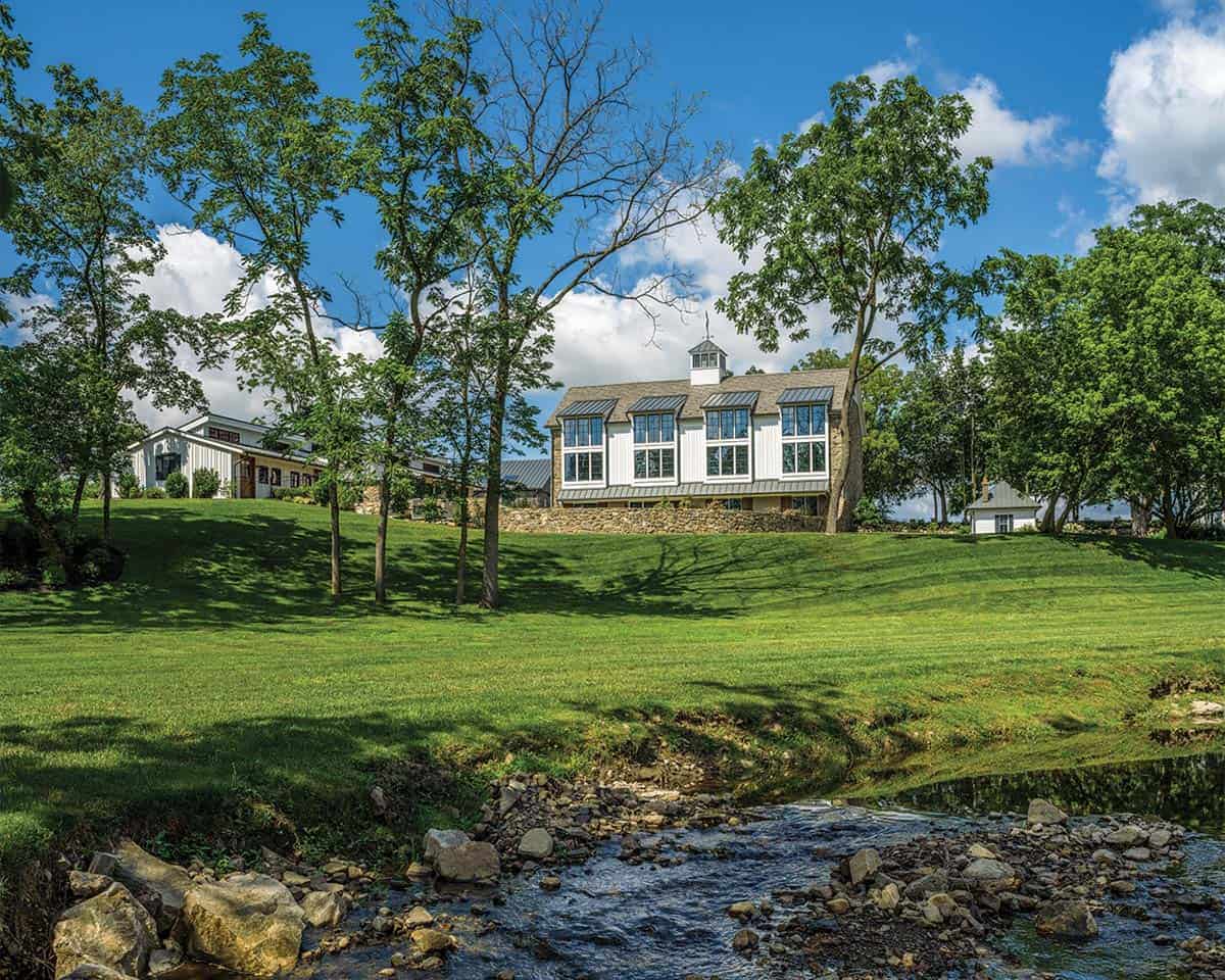 Historic Pennsylvania bank barn conversion viewed across a rocky creek, showing white board-and-batten upper story with stone foundation on a lush green hillside