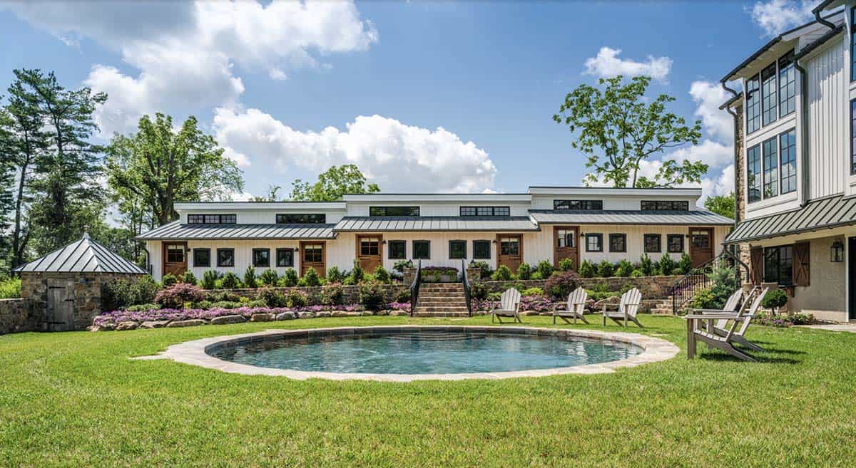 Rear view of converted barn home with circular pool, Adirondack chairs, white board-and-batten guest wing, and terraced stone retaining walls with flowering shrubs
