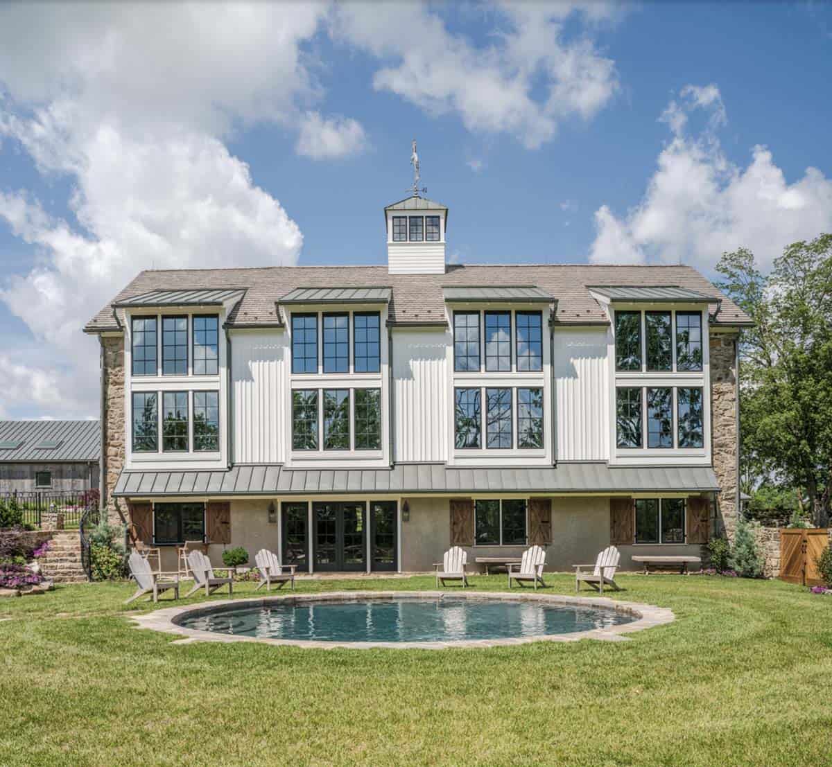 Back facade of converted bank barn featuring floor-to-ceiling multi-pane windows, white vertical siding, original stone lower level, cupola with weather vane, and circular pool