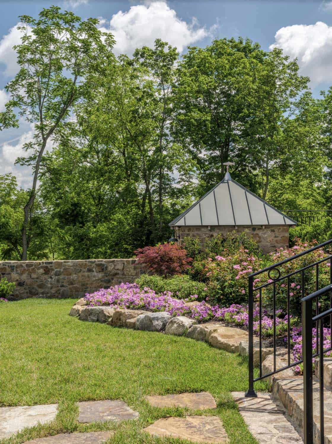 Stone garden folly with pyramid metal roof surrounded by purple azaleas, stone retaining wall, flagstone path, and black iron stair railing