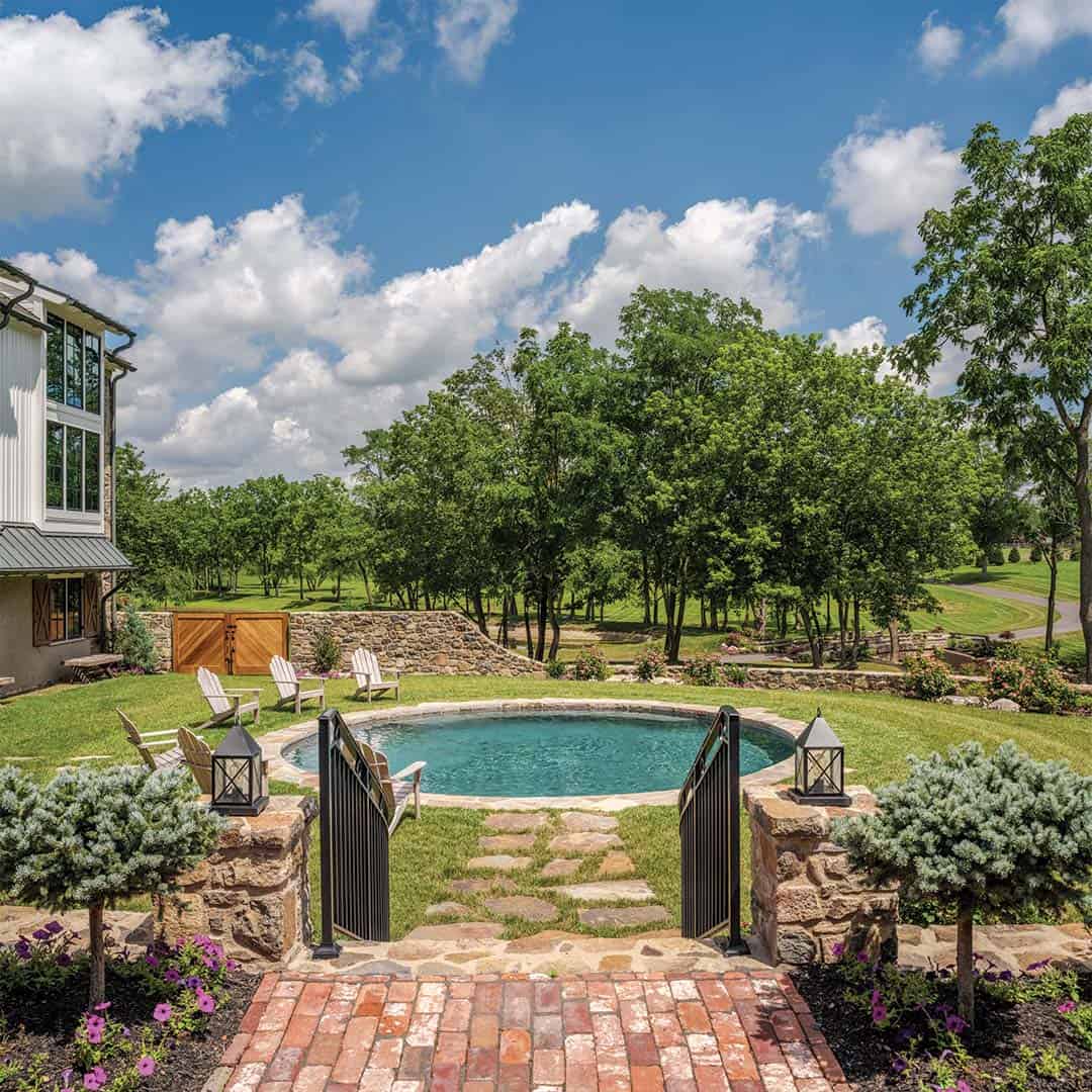 Aerial view of circular plunge pool with stone surround, brick pathway entry gate with black iron railings, Adirondack chairs, and mature orchard trees beyond stone wall