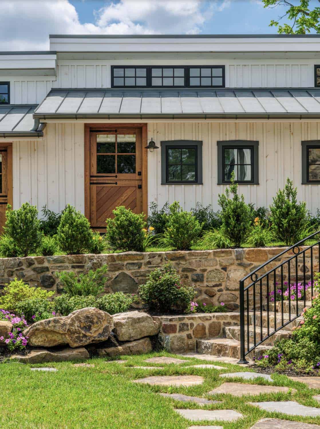 White board-and-batten carriage house wing with warm wood Dutch door, black-trimmed windows, metal roof, and terraced stone landscape beds with flowering plants