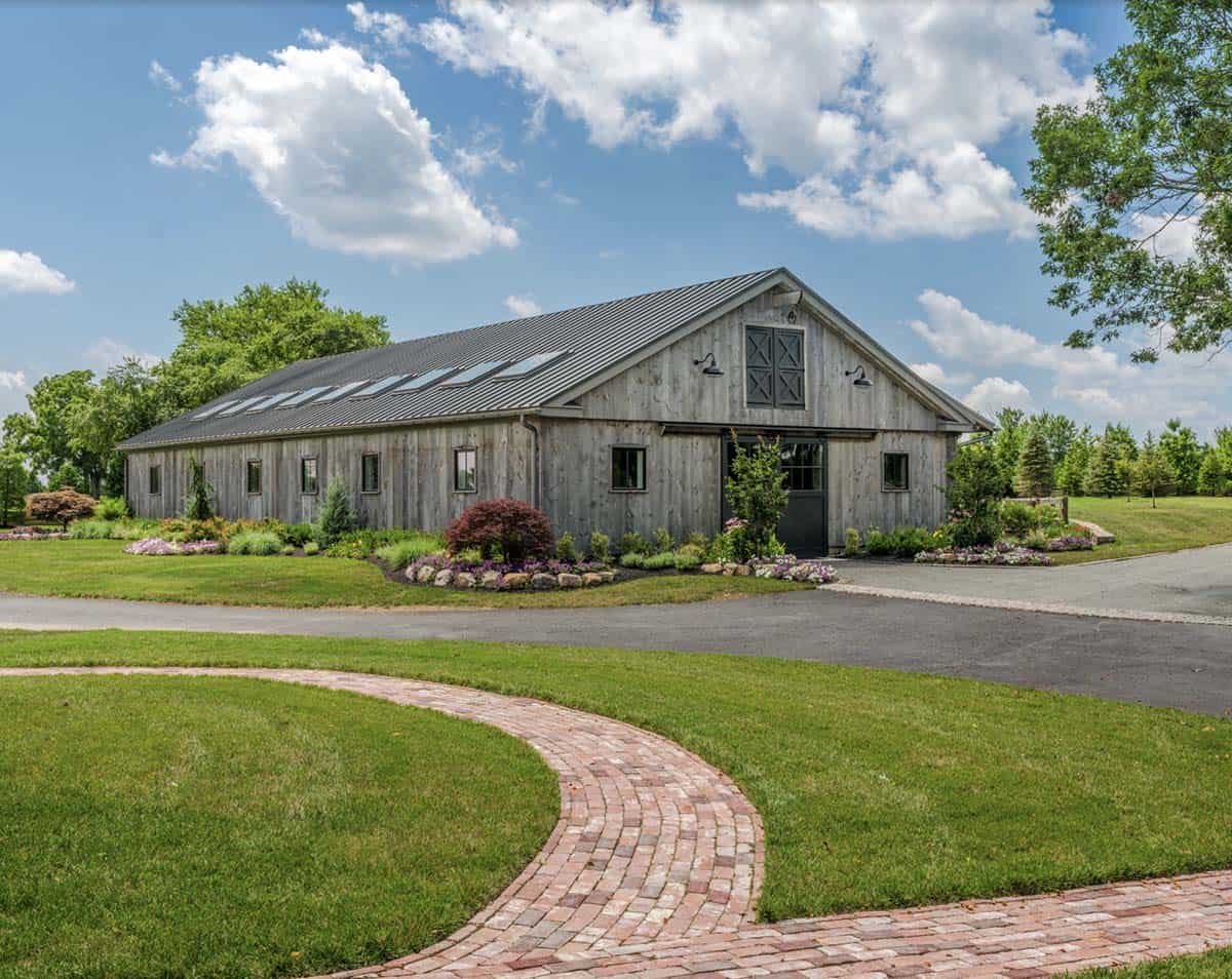 Weathered gray board-and-batten new barn structure with standing-seam metal roof and skylights, gable-end X-pattern shutters, gooseneck barn lights, and curved brick pathway