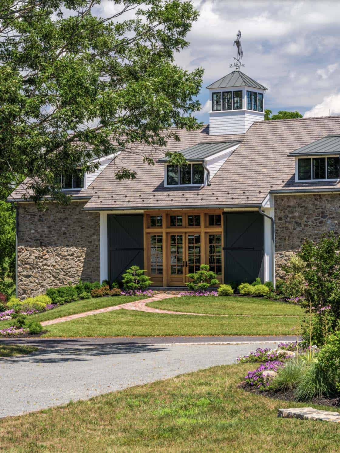 Close-up of historic bank barn entry facade showing original fieldstone walls, oversized warm wood French doors flanked by sliding charcoal barn doors, cupola with horse weather vane, and cedar shake roof