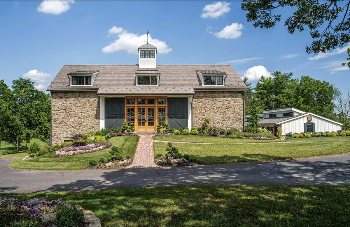 Front view of converted fieldstone bank barn with cedar shake roof, dormer windows, cupola weather vane, warm wood French door entry, and brick pathway through manicured landscaping