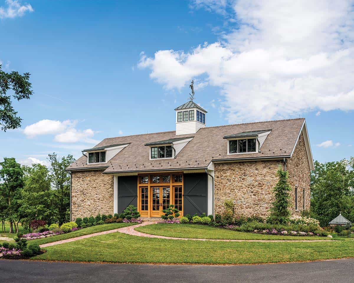 view of converted stone bank barn showing cedar shake roof, eyebrow dormers, cupola with horse weather vane, charcoal sliding barn doors, and flowering garden beds