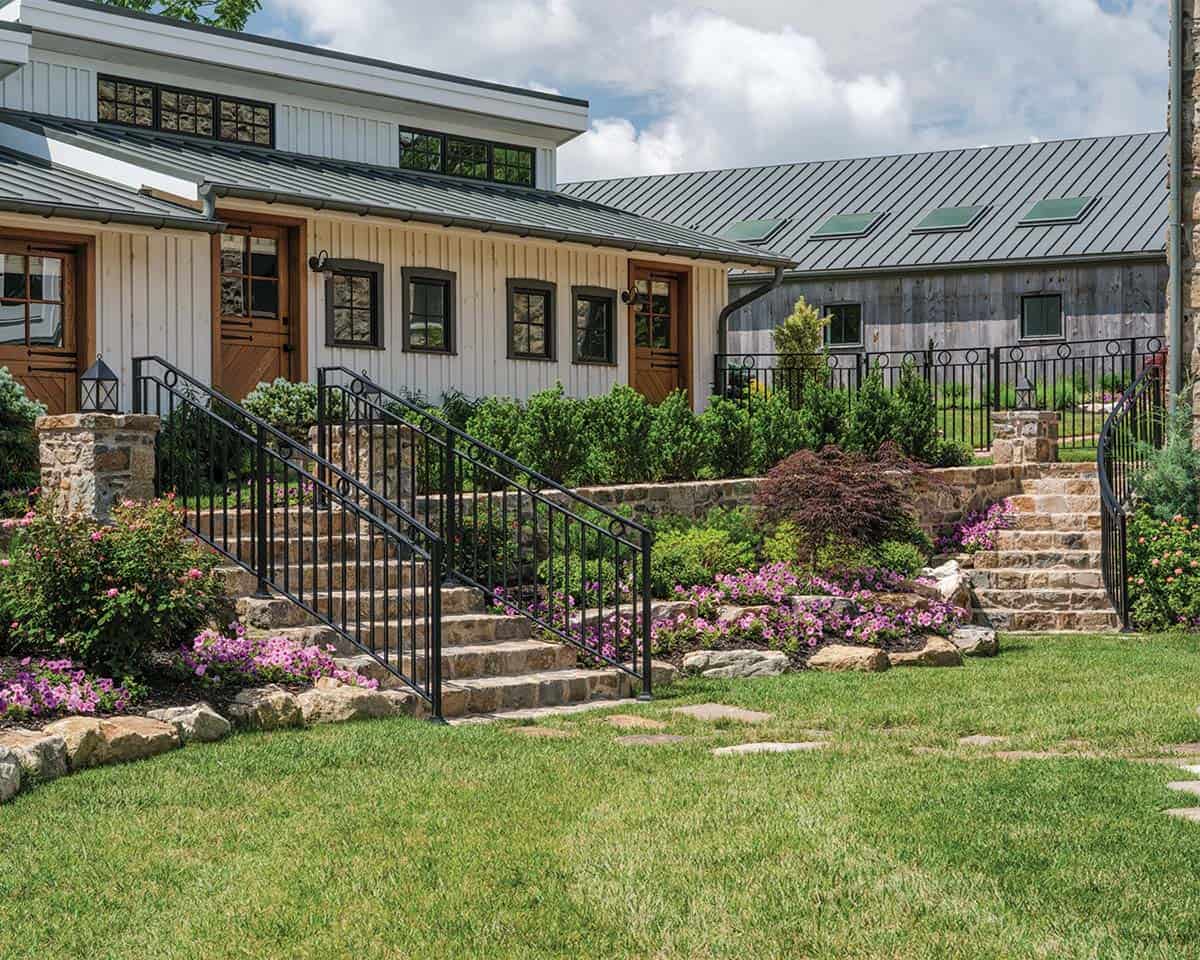 Tiered stone steps with black iron railings leading up to white board-and-batten guest wing, with colorful flower borders, Japanese maple, and weathered gray barn structure beyond