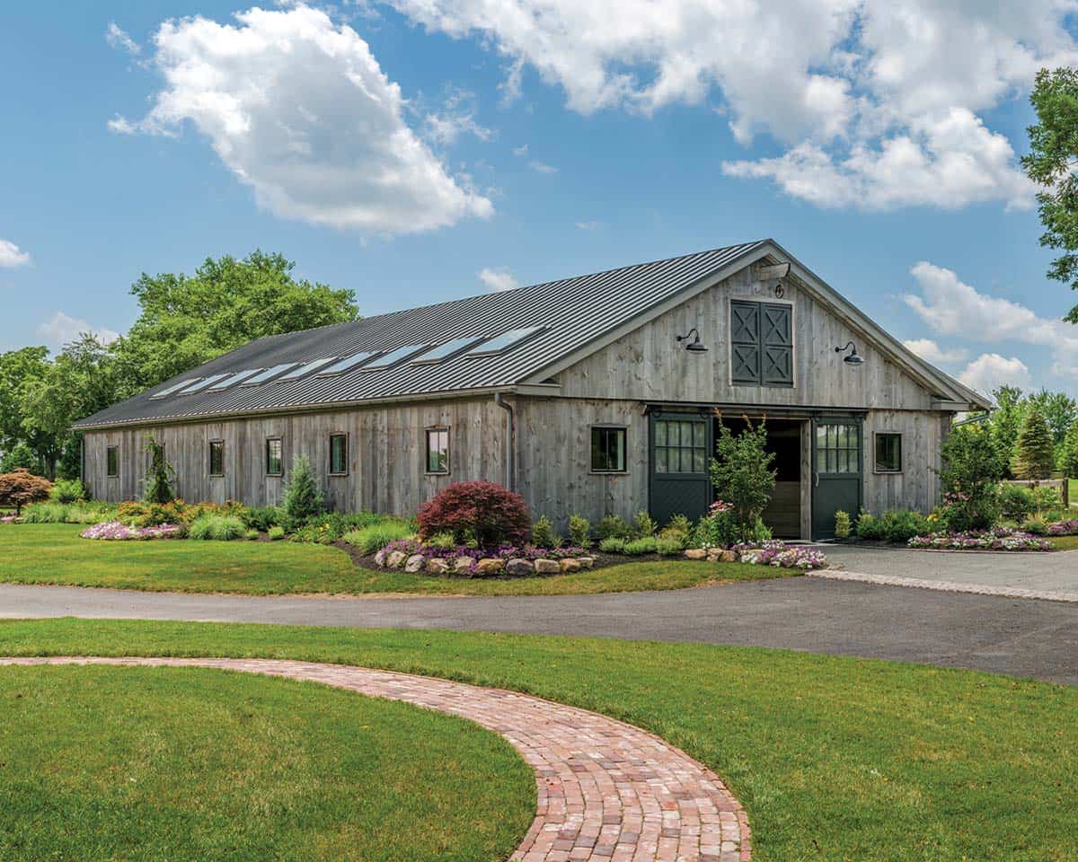 Side and front view of new weathered gray board-and-batten barn with standing-seam metal roof, roof skylights, X-pattern gable shutters, gooseneck barn lights, and curved brick driveway path