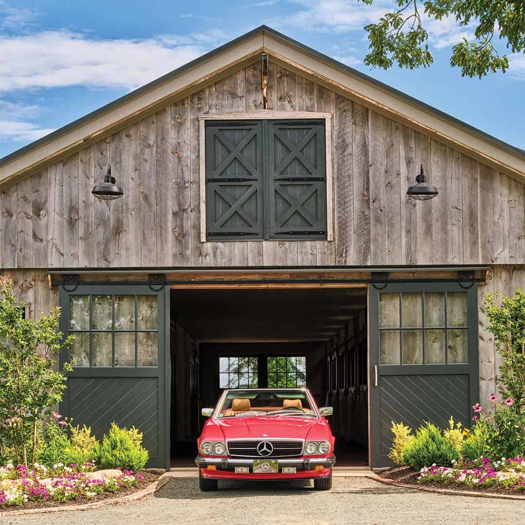 Weathered gray barn gable end with charcoal X-pattern loft doors, gooseneck barn lights, and large sliding carriage doors open to reveal a vintage red Mercedes-Benz convertible inside