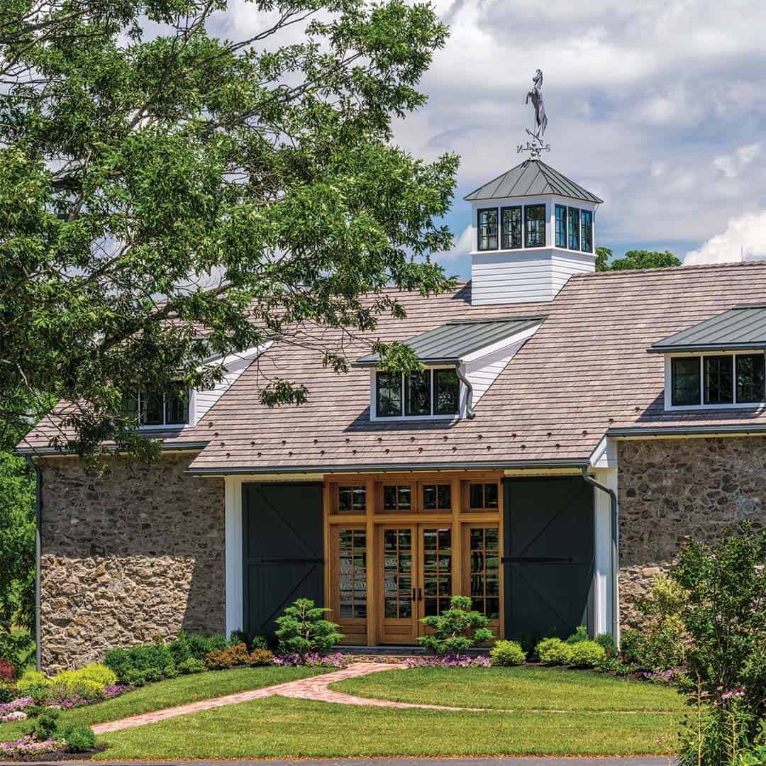 Close-up of converted fieldstone bank barn entry with warm wood French doors, flanking charcoal sliding barn doors, cedar shake roof, eyebrow dormers, and cupola with horse weather vane
