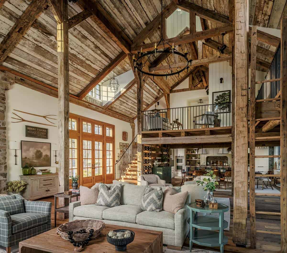 Soaring open-plan barn conversion interior with original exposed timber frame, reclaimed wood plank ceiling, iron chandelier, loft with black railing, and open kitchen beyond the living area