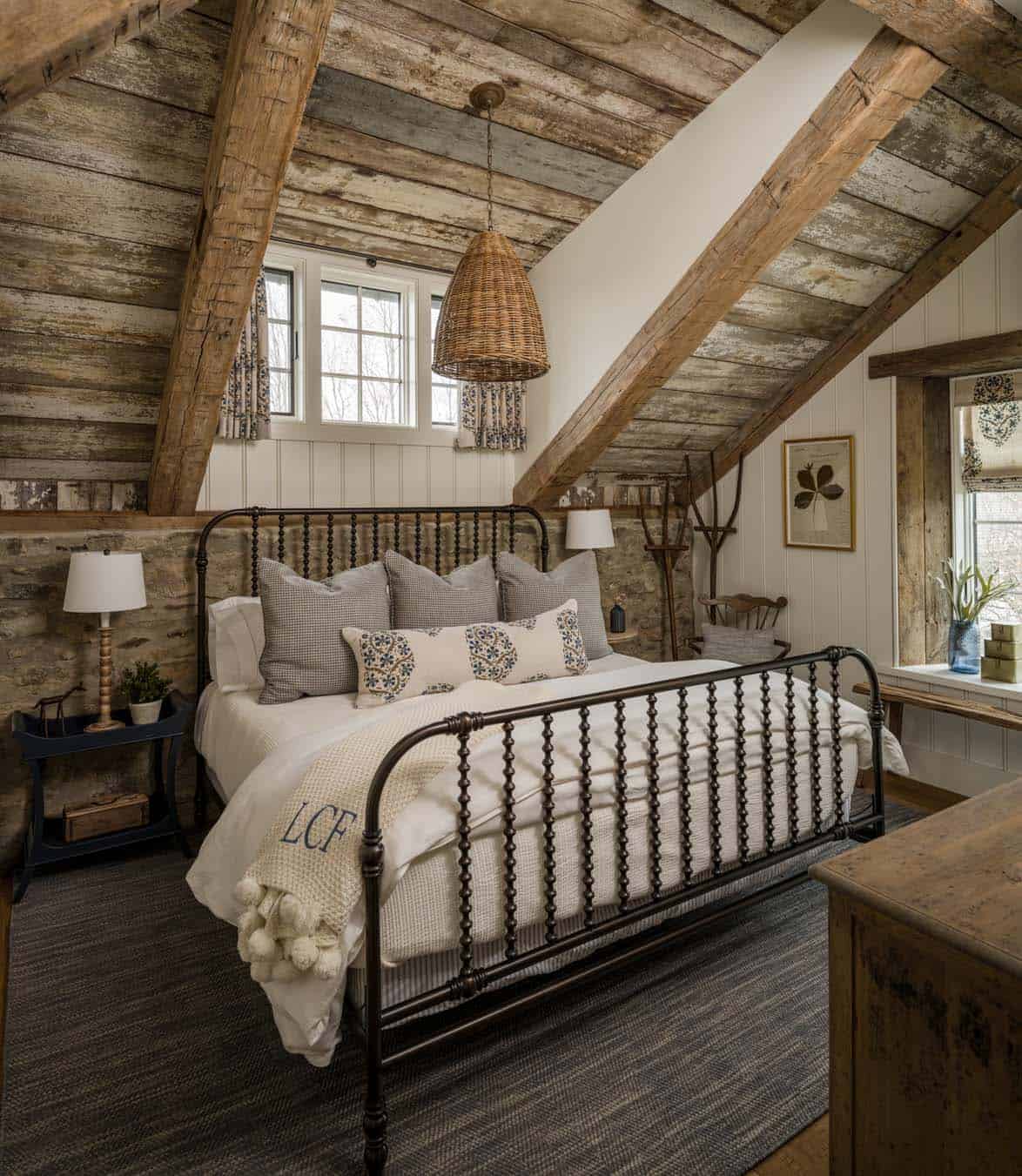 Rustic barn conversion bedroom with original reclaimed wood plank ceiling, diagonal timber beams, dark iron spindle bed, wicker pendant light, original stone wall, and blue-and-white patterned bedding