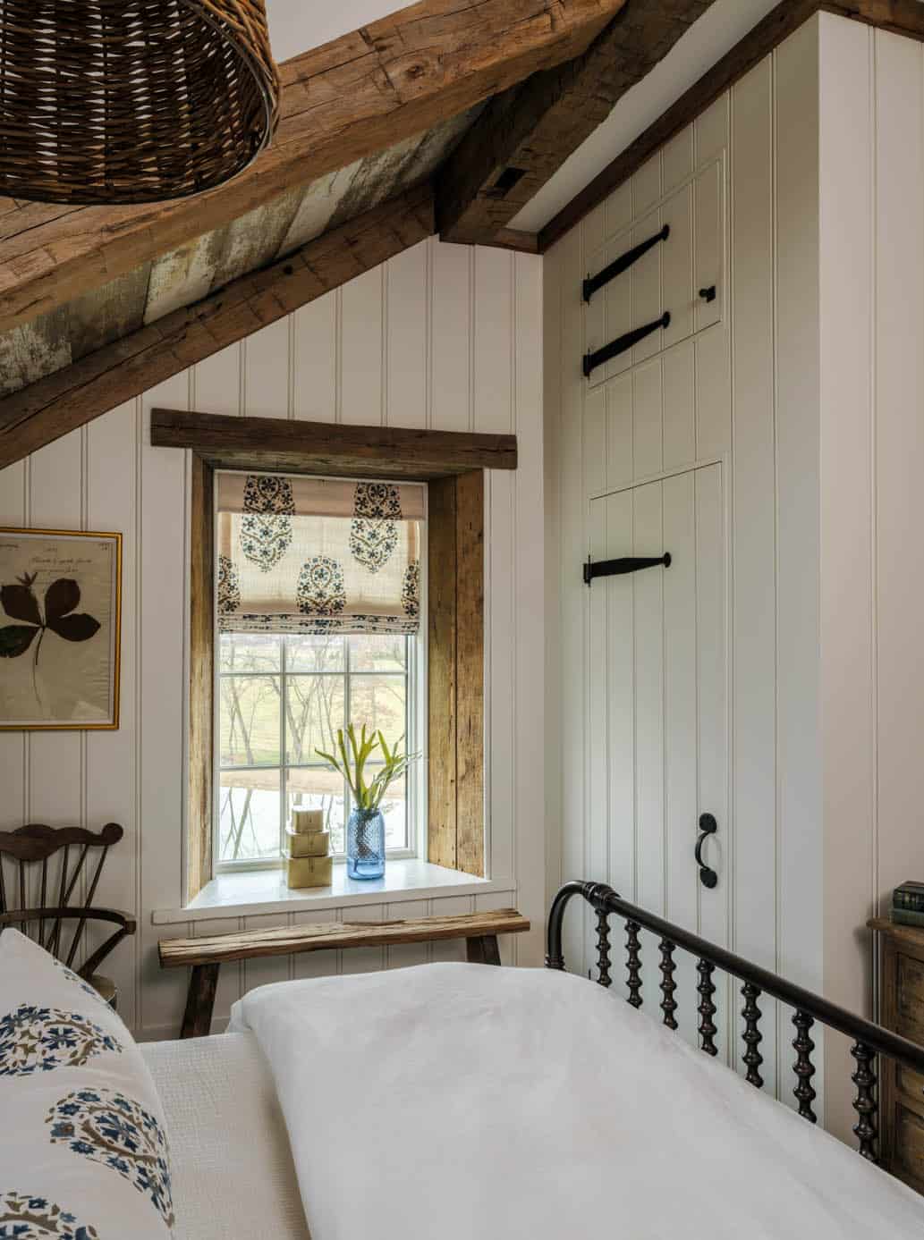 Bedroom detail showing reclaimed wood beam framing a grid-pane window with blue floral Roman shade, white beadboard walls, a built-in painted cabinet with black iron hardware, and rustic wood bench below the sill