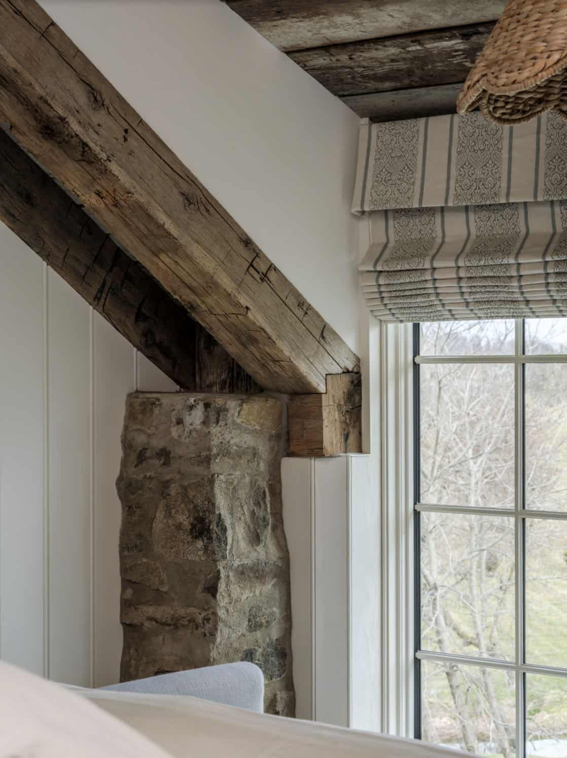 Close-up architectural detail of original fieldstone wall and weathered timber beam meeting at a bedroom window corner with striped Roman shade and view to bare winter trees