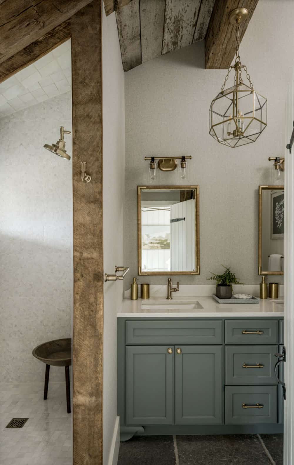 Barn conversion bathroom with sage green double vanity, brass fixtures, gold geometric pendant lantern, original reclaimed timber beam, open frameless marble-tile shower, and slate floor tile