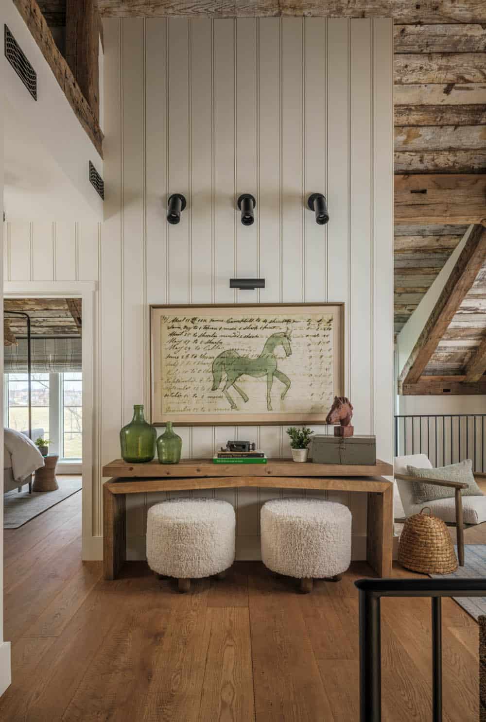 Upper hallway landing with white beadboard walls, framed vintage horse ledger artwork lit by three black picture lights, rustic wood console table with two shearling ottomans beneath, and exposed timber ceiling