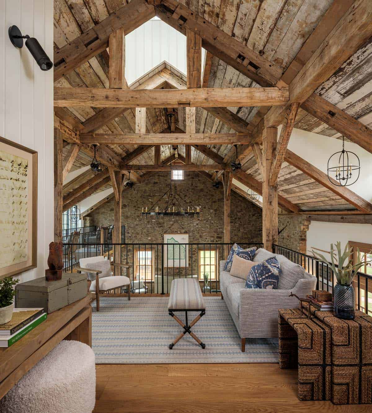 Upper mezzanine loft sitting area with dramatic exposed original timber truss framework, reclaimed plank ceiling, original stone end wall, gray sofa with blue pillows, iron oval pendant, and mezzanine railing overlooking lower level
