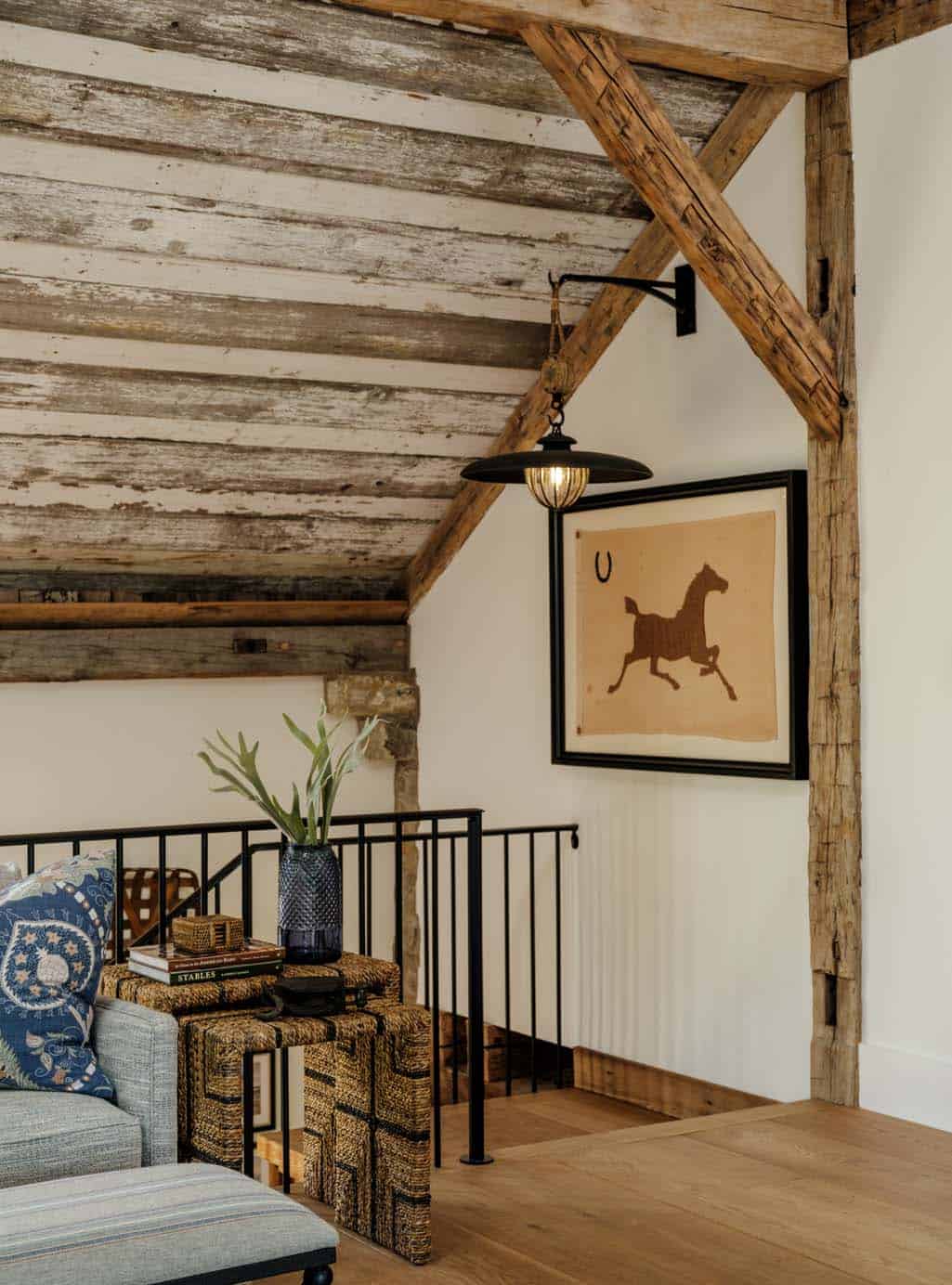 Loft mezzanine corner detail with original timber knee brace, reclaimed plank ceiling, vintage barn pulley pendant light, framed horse silhouette artwork, woven seagrass cube ottomans, and black iron railing