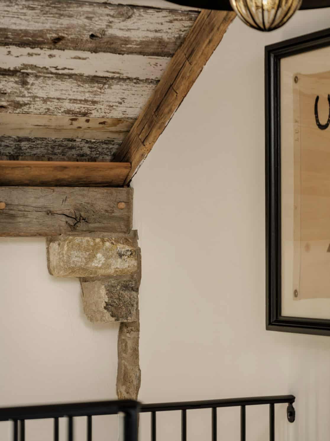 Close-up architectural detail of original hand-hewn timber rafter tail sitting on a rough-cut fieldstone corbel against white plaster wall, with a partial view of a framed horse print