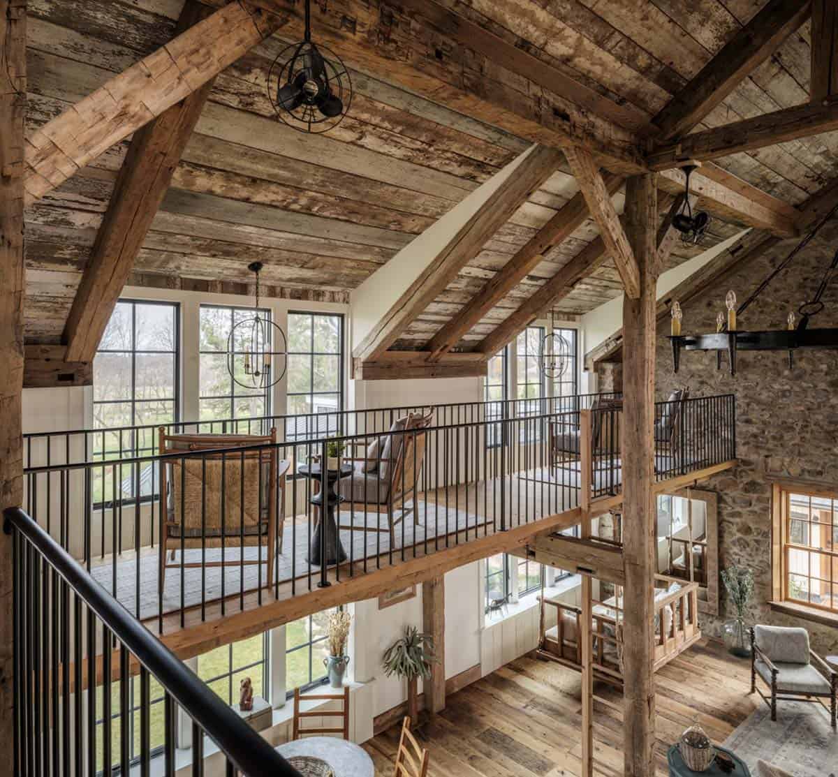 Looking down from upper stair landing into the multi-story barn home interior showing iron mezzanine railing, original heavy timber frame, reclaimed plank ceiling, vintage cage fan pendants, large grid-pane windows, and open living area below