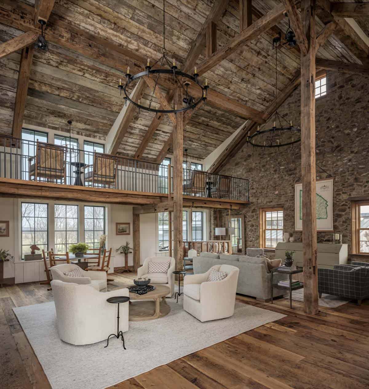 Dramatic double-height barn home living room with original timber frame, reclaimed plank ceiling, two iron wagon wheel chandeliers, mezzanine balcony with iron railing, original fieldstone end wall, and white boucle seating grouping