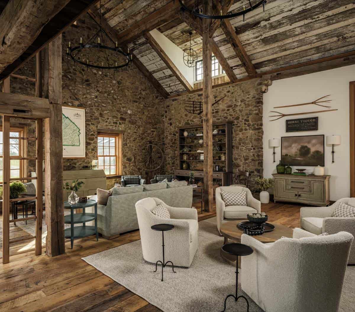 Barn conversion great room showing original fieldstone wall, reclaimed timber posts, iron chandelier, built-in dark wood bookcase styled with antiques, rope swing, vintage spinning wheel, and cream upholstered seating