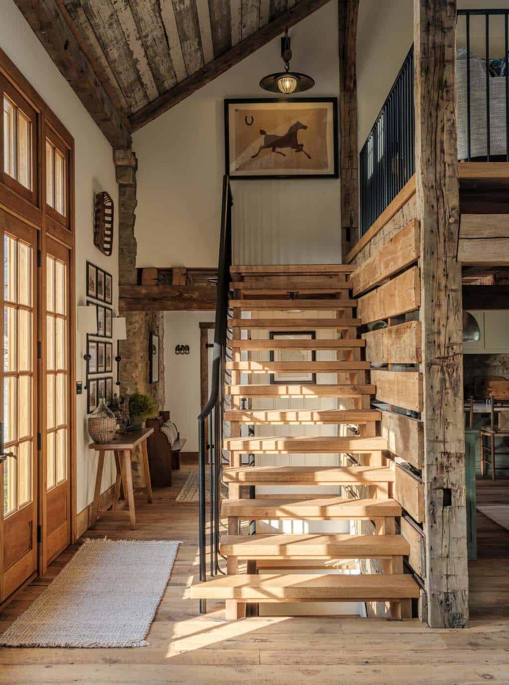 Open-tread floating wood staircase with black iron railing rising to loft level beside warm wood French entry doors, reclaimed plank ceiling, original timber beams, and framed horse artwork above landing