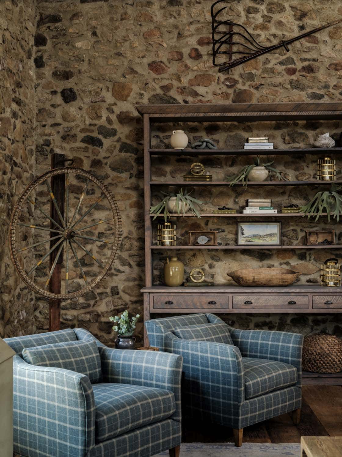 Original fieldstone interior wall with large dark wood open bookcase styled with antiques, pottery, plants, and a framed landscape painting, flanked by two blue plaid upholstered armchairs and a vintage spinning wheel