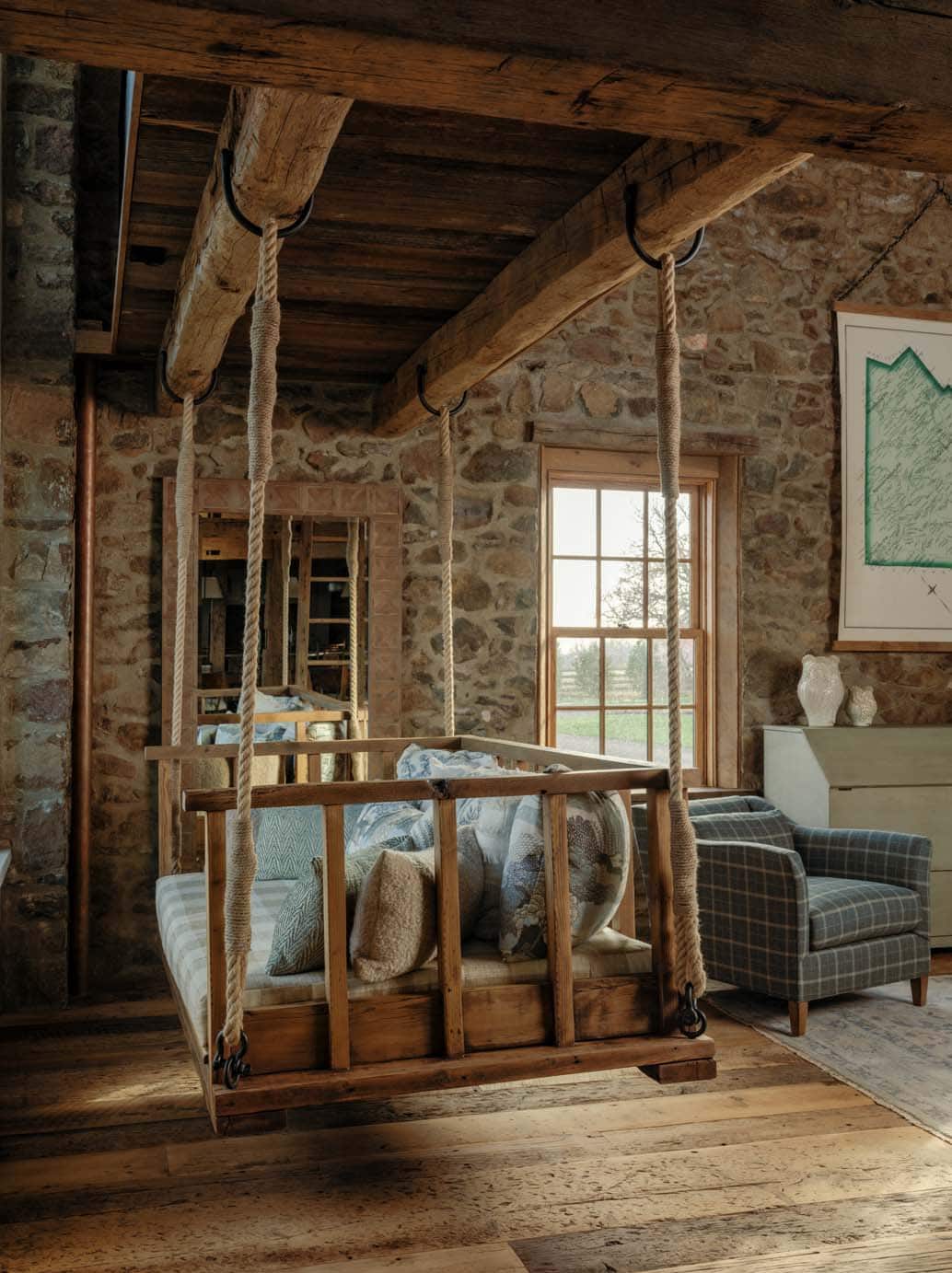 Rustic wooden rope-hung indoor porch swing loaded with cushions and pillows suspended from original heavy timber beams against original fieldstone wall beside a grid-pane window with pastoral views