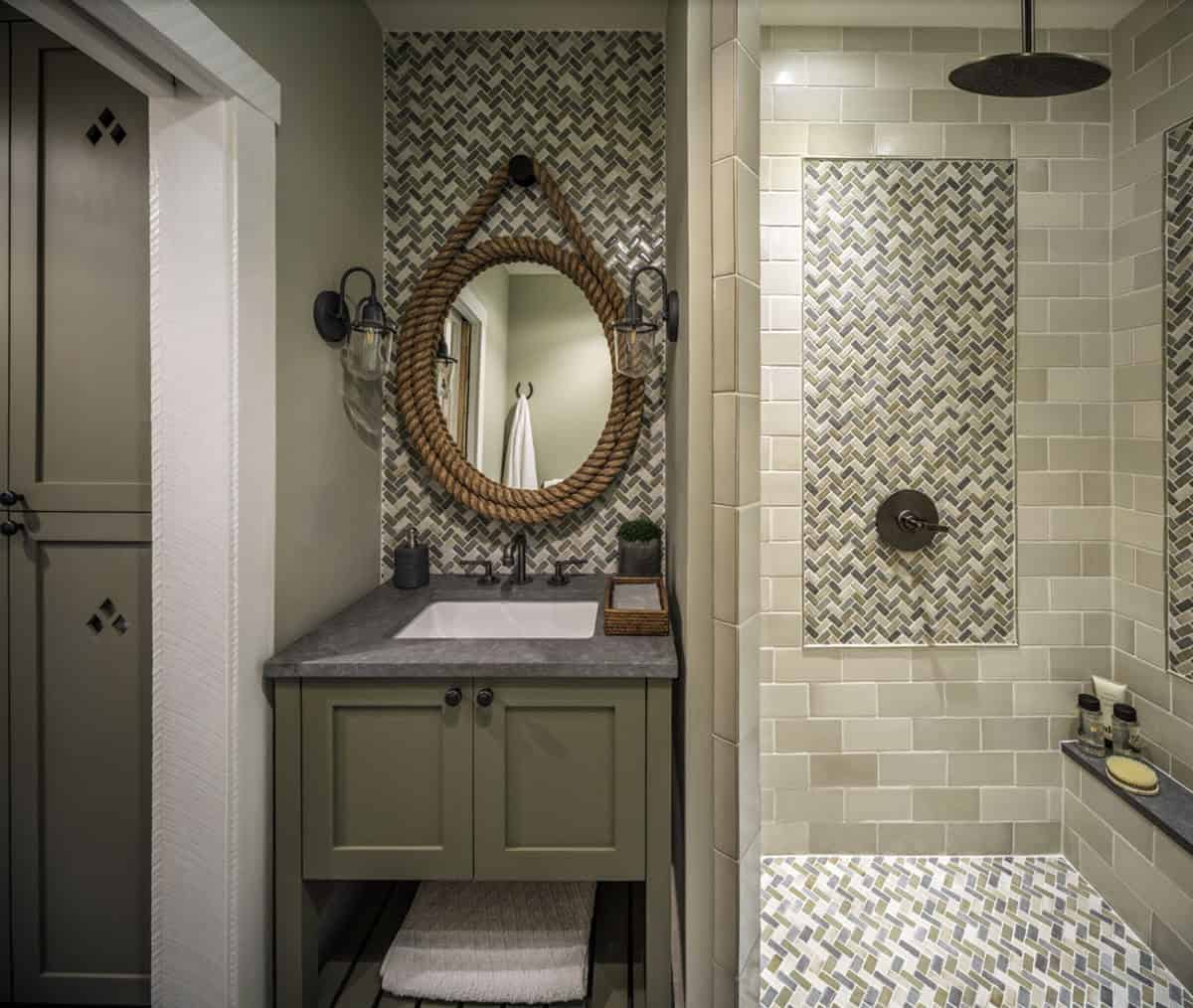 Barn conversion guest bathroom with sage green vanity cabinet, concrete countertop, rope-wrapped round mirror, black wall sconces, herringbone mosaic tile accent wall, and open subway tile walk-in shower with rain head