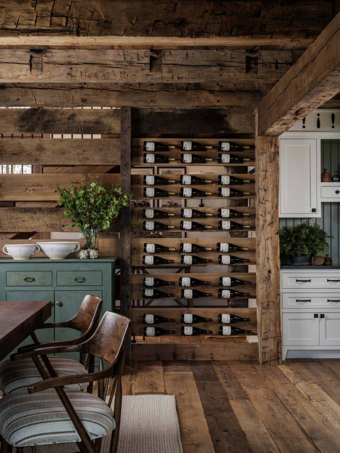 Barn home dining area wine storage wall built into original reclaimed timber structural frame showing rows of wine bottles displayed horizontally, green painted sideboard beneath, and white kitchen cabinetry beyond