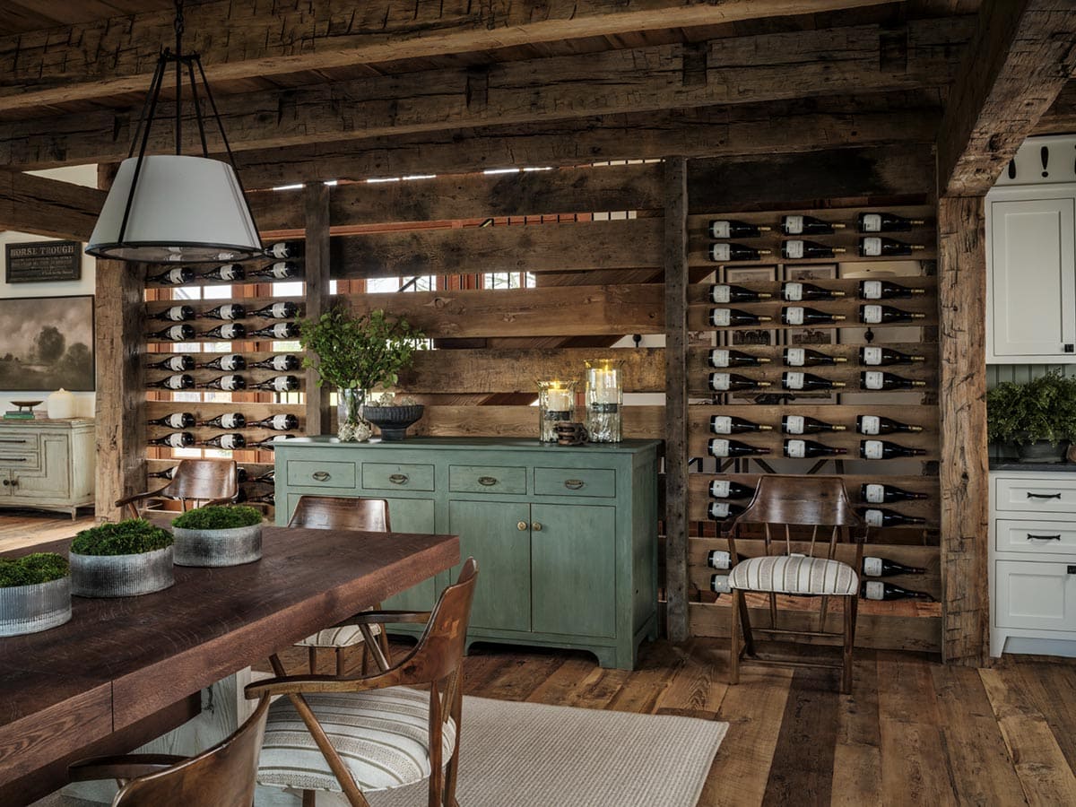 Dining room with large dark wood trestle table, mixed wood and striped upholstered chairs, three white cone pendant lights, reclaimed timber ceiling, and wine bottle display wall built into original timber frame