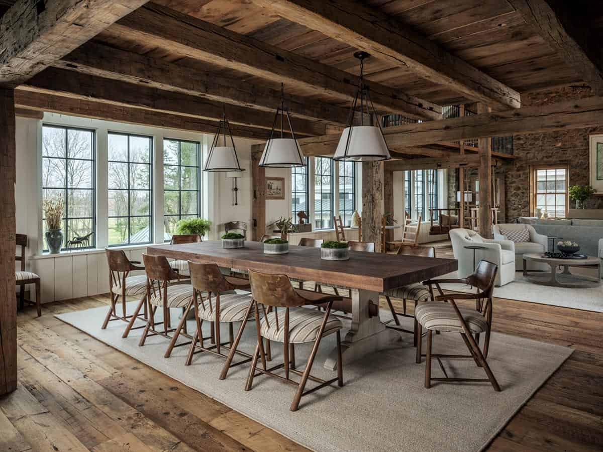 Expansive barn home dining room with long dark wood trestle table seating twelve, three white cone pendants, original heavy timber beams, reclaimed plank ceiling, large grid-pane windows, and open view to living area