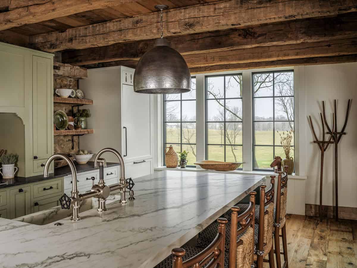 Barn home kitchen with large marble waterfall island, bridge faucet with industrial wheel handles, hammered metal dome pendant, sage green cabinetry with black hardware, reclaimed open shelving on original stone wall, and grid-pane windows with country views