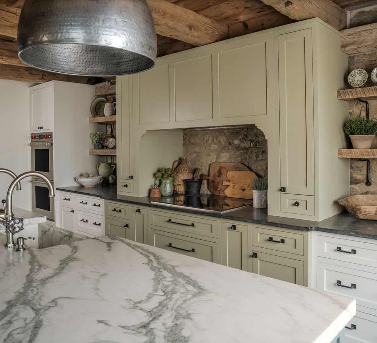 Barn conversion kitchen showing sage green shaker cabinetry with black hardware, arched cooktop alcove with original stone backsplash, hammered metal dome pendant, marble island countertop, and reclaimed timber ceiling beams