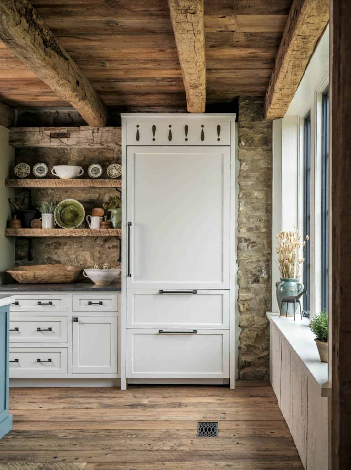 Barn kitchen corner featuring white panel-front refrigerator with decorative cutout crown detail, reclaimed timber open shelves on original stone wall displaying pottery and dishes, and white lower cabinets with black hardware