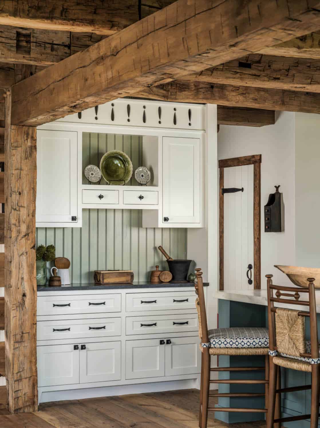 Kitchen hutch area with white cabinetry, sage green beadboard backsplash, decorative cutout crown molding, dark soapstone countertop, original heavy timber frame surround, and rush-seat bar stools at the island beyond