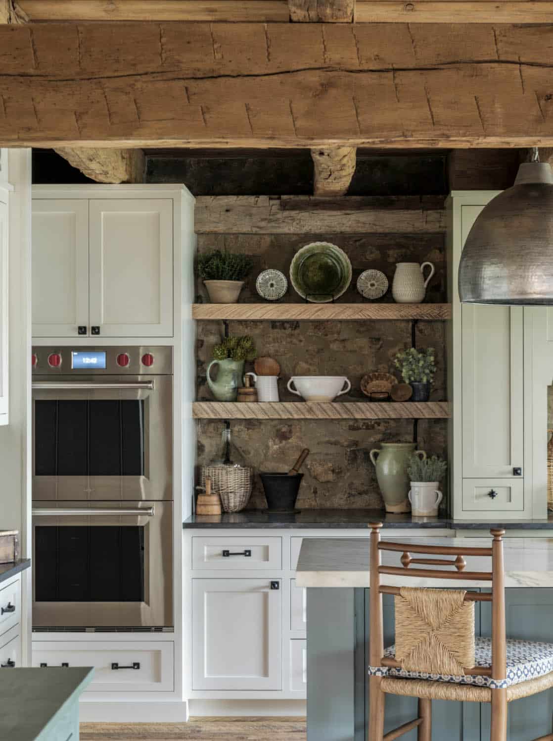 Barn kitchen wall detail showing stainless double wall oven beside reclaimed timber open shelves on original fieldstone backsplash, styled with green pottery, white pitchers, and woven baskets under a hammered metal dome pendant