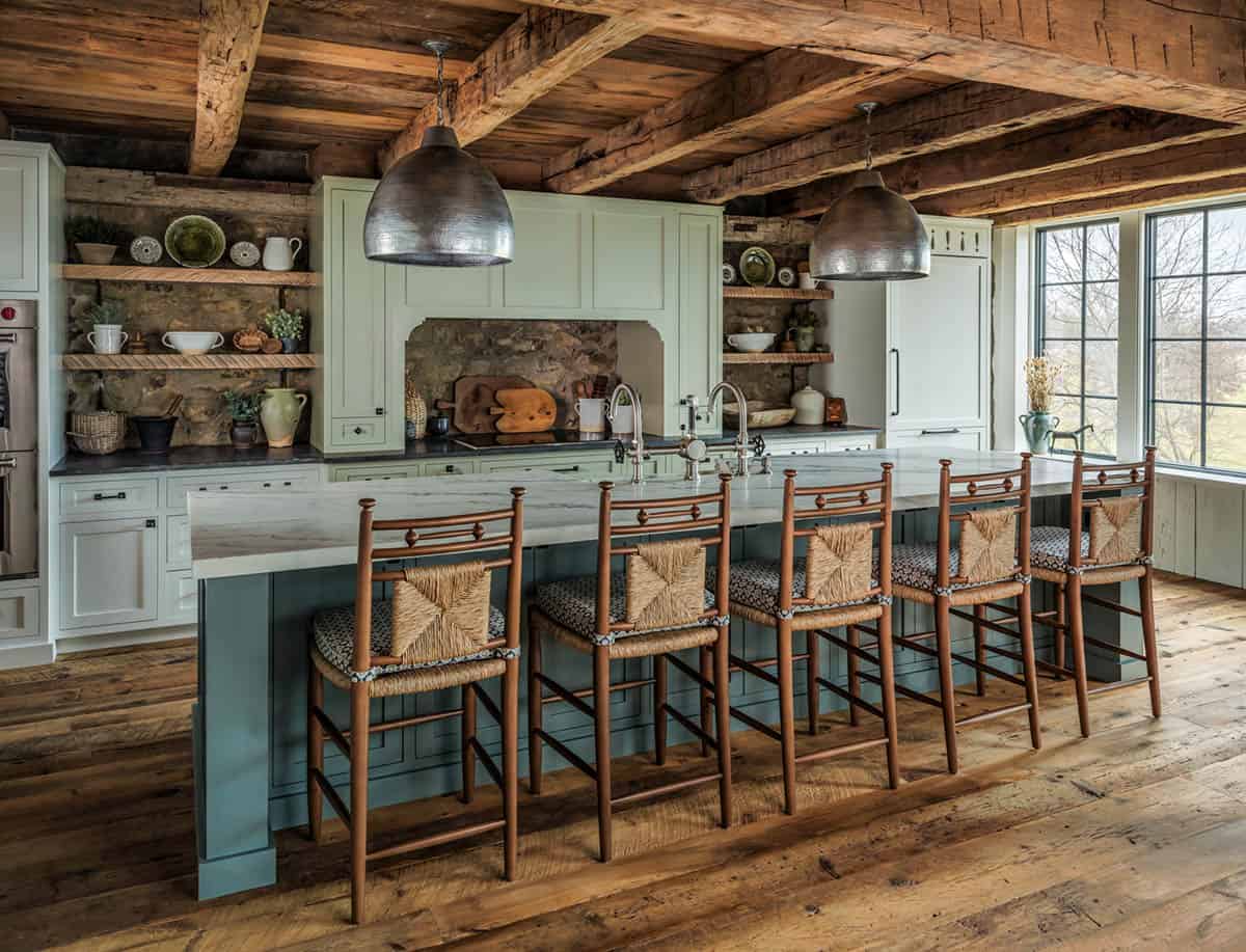 barn home kitchen view with long teal-green island seating six rush-seat bar stools, sage green cabinetry, twin hammered metal dome pendants, reclaimed timber ceiling, stone backsplash cooktop alcove, and large grid-pane windows